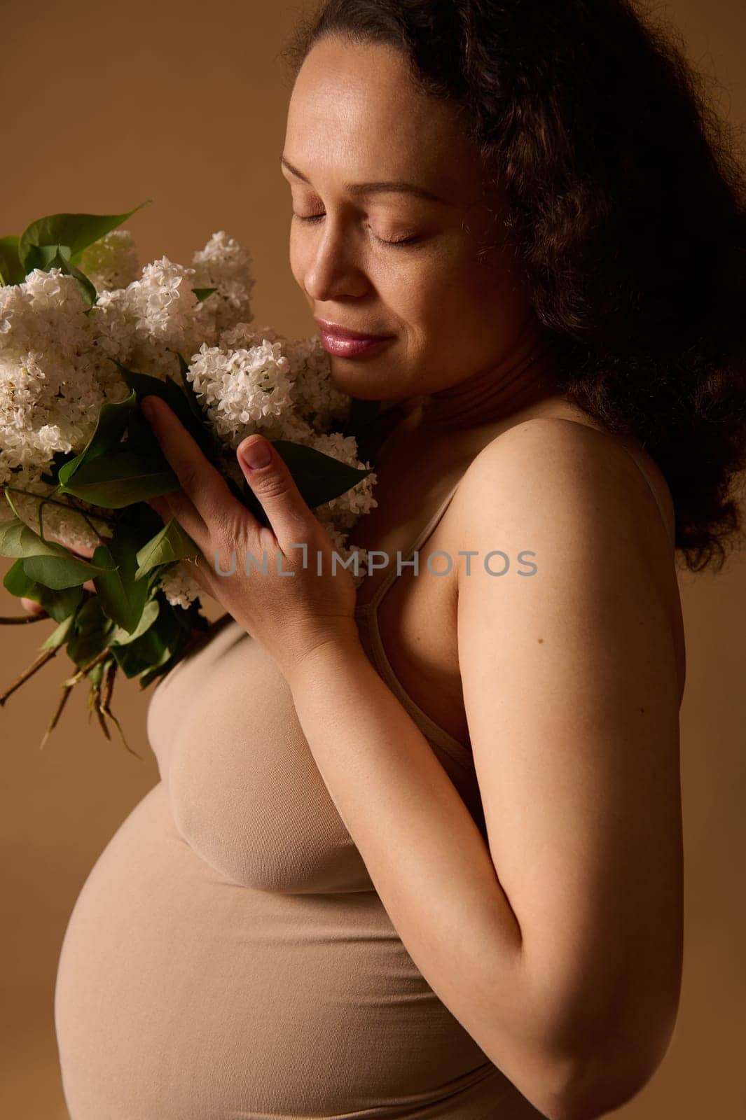 Close-up pretty pregnant woman with closed eyes, in beige lingerie, holding bunch of lilacs. Pregnancy. Expecting a baby by artgf