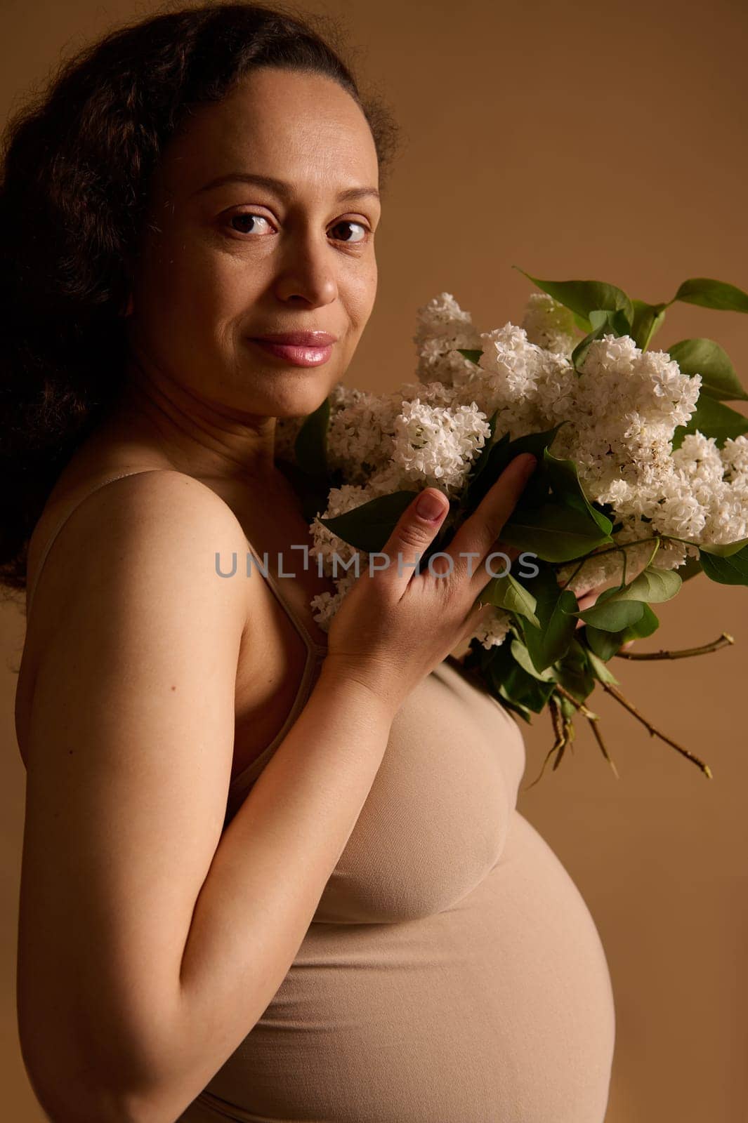Attractive multiethnic gravid woman, pregnant expectant mother in beige lingerie, holding bouquet of blooming lilacs, isolated studio background. Pregnancy 28 week Fertility Gynecology Women's health