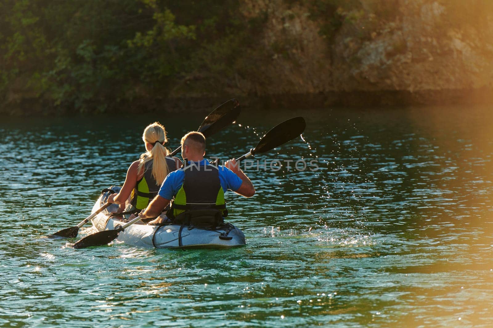 A young couple enjoying an idyllic kayak ride in the middle of a beautiful river surrounded by forest greenery in sunset time.