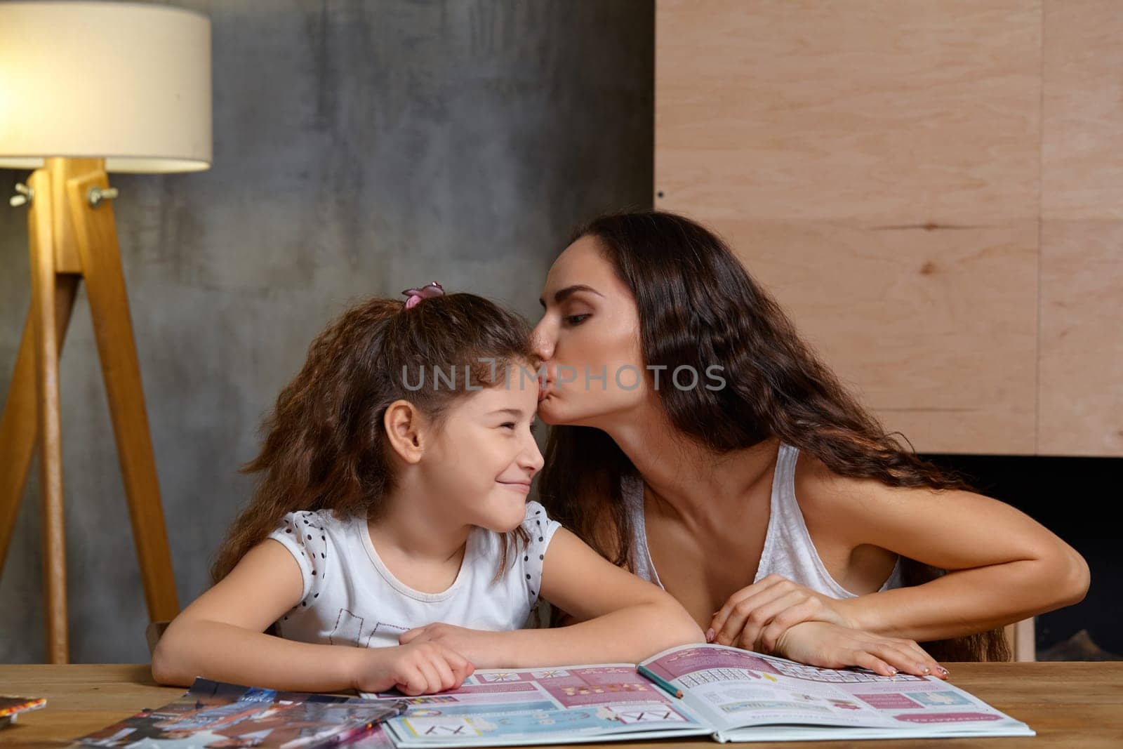 Portrait of a mother helping her small sweet and cute daughter to make her homework indoors. Mom is kissing her daughter.
