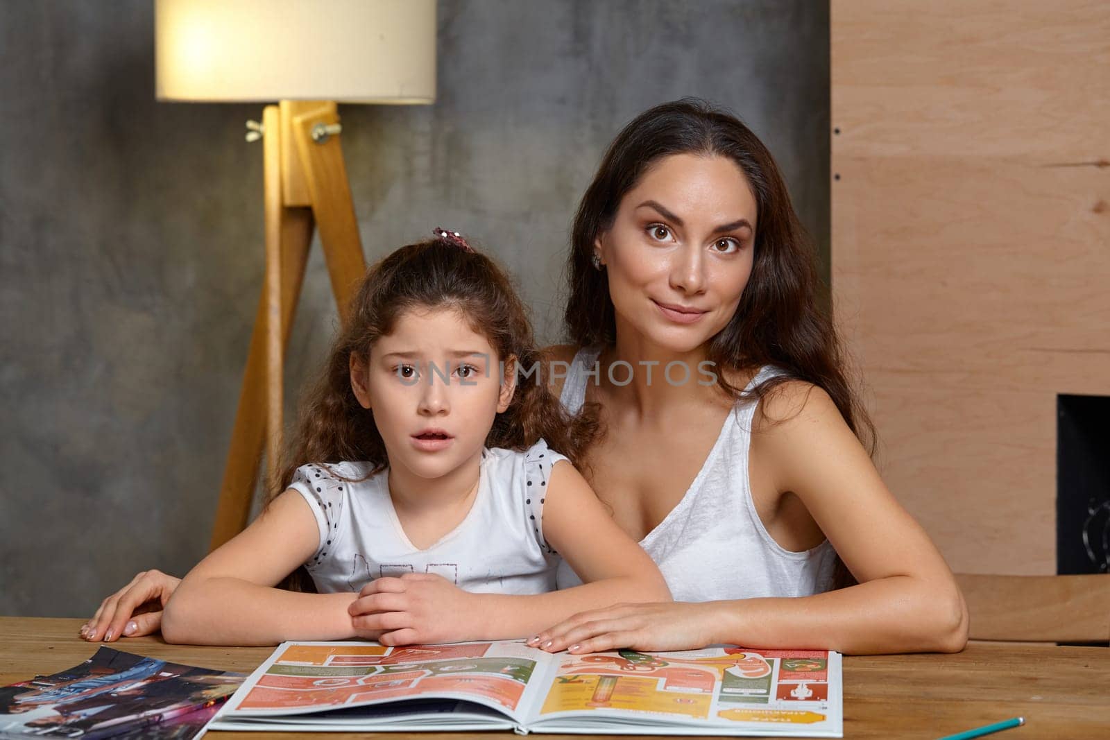 Portrait of a mother helping her small sweet and cute daughter to make her homework indoors. They are looking at the camera.