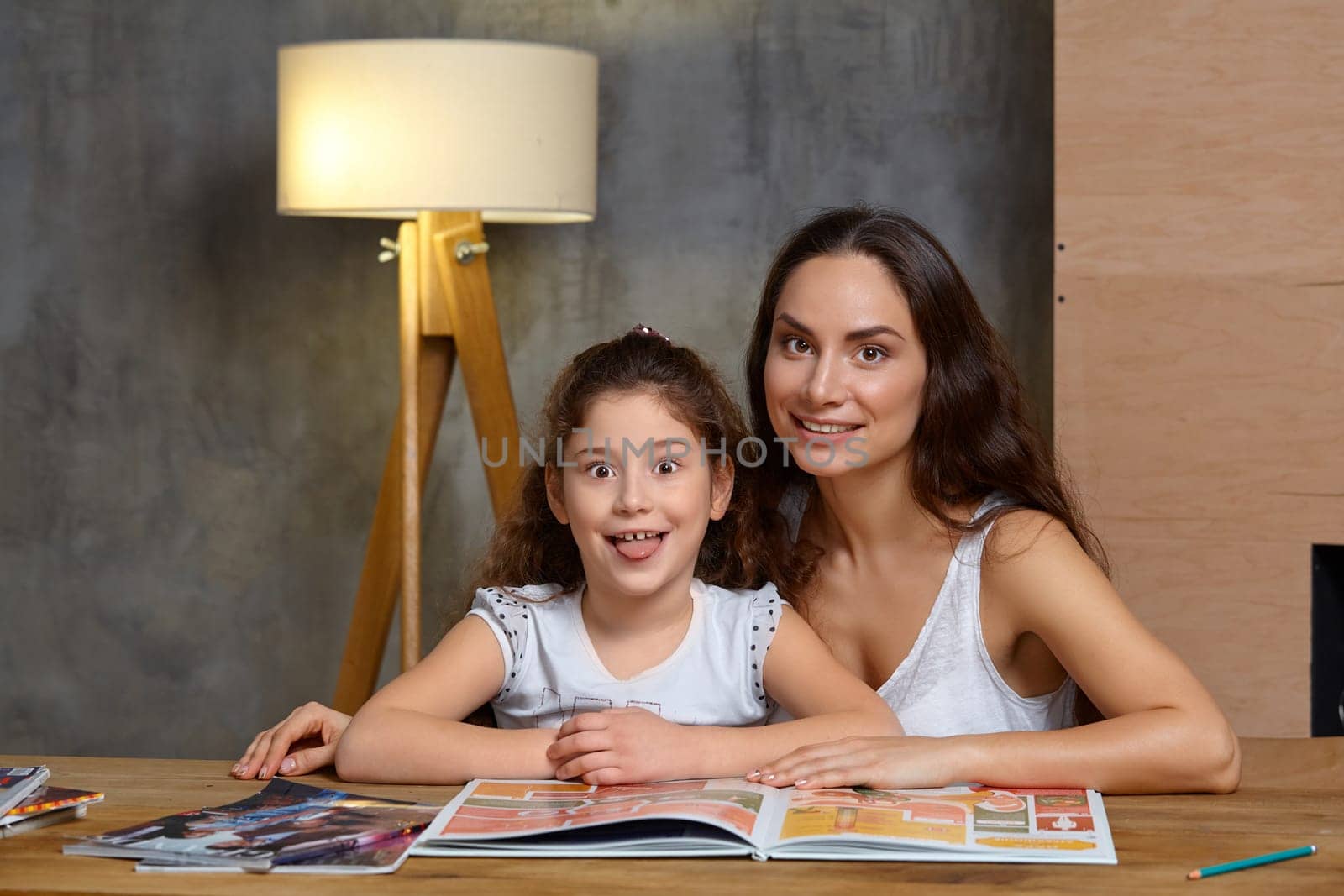 Portrait of a mother helping her small sweet and cute daughter to make her homework indoors. They are looking at the camera. Mom is smiling and her daughter is showing her tongue.
