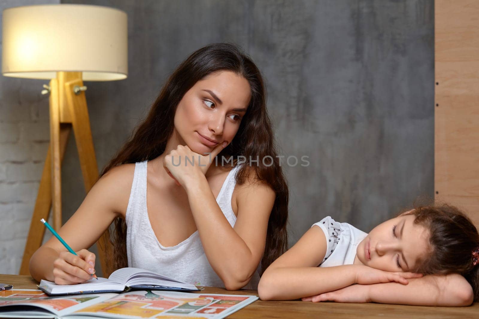 Portrait of a mother helping her small sweet and cute daughter to make her homework indoors. Mom is looking at her daughter holding a pencil in her hand, daughter is sleeping.