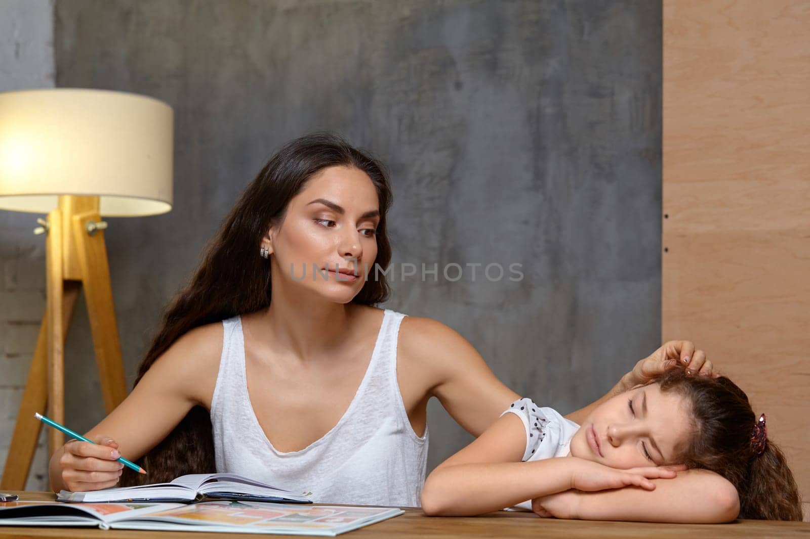 Portrait of a mother helping her small sweet and cute daughter to make her homework indoors. Mom is stroking her daughter holding a pencil in her hand, daughter is sleeping.