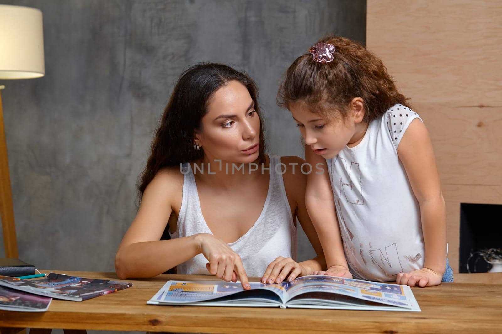 Portrait of a mother helping her small and cute daughter to make her homework indoors. Mom is showing something in the notebook and her daughter is looking at it.
