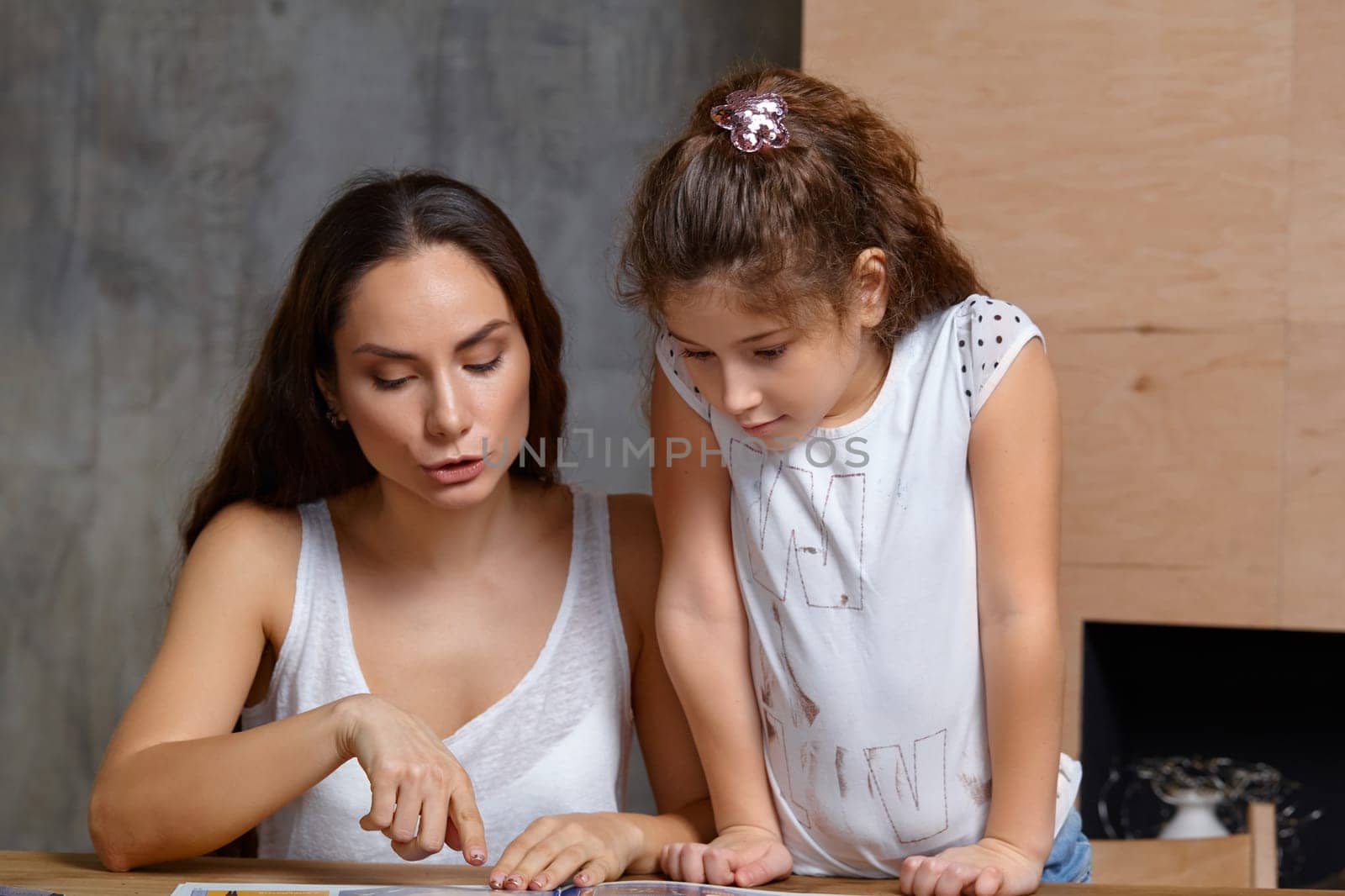 Portrait of a mother helping her small sweet and cute daughter to make her homework indoors. Mom is showing something in the notebook and her daughter is looking at it.