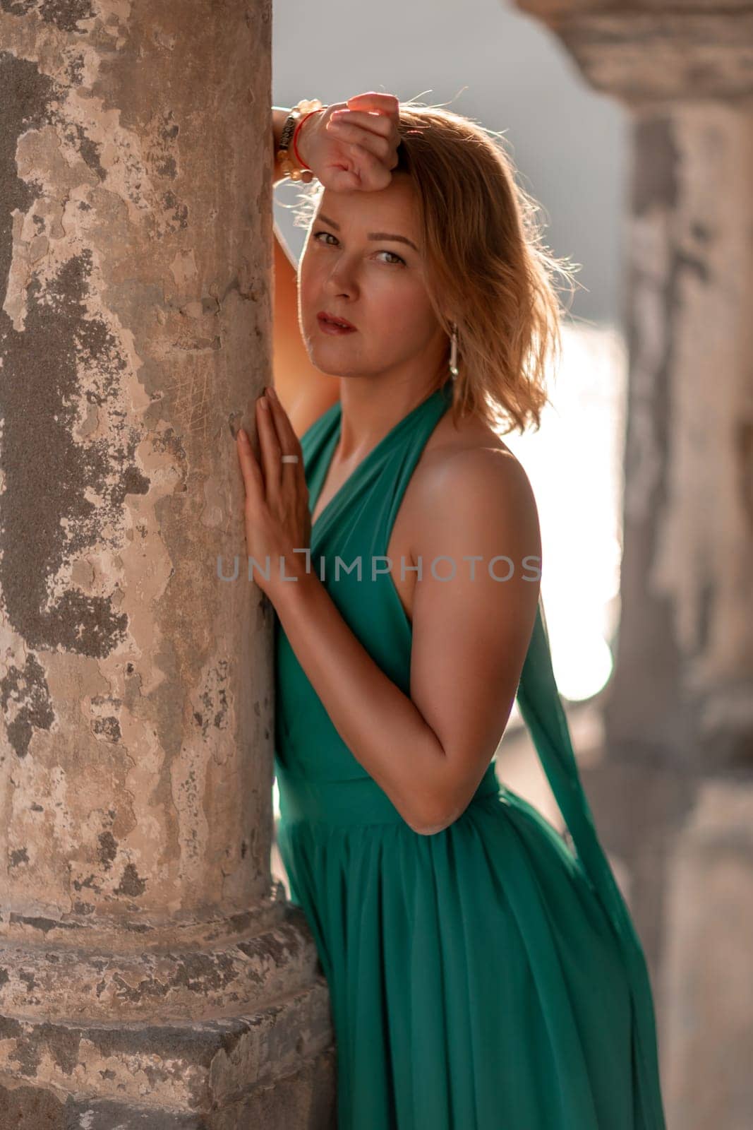 Happy blonde in a long mint dress posing against the backdrop of the sea in an old building with columns. Girl in nature against the blue sky