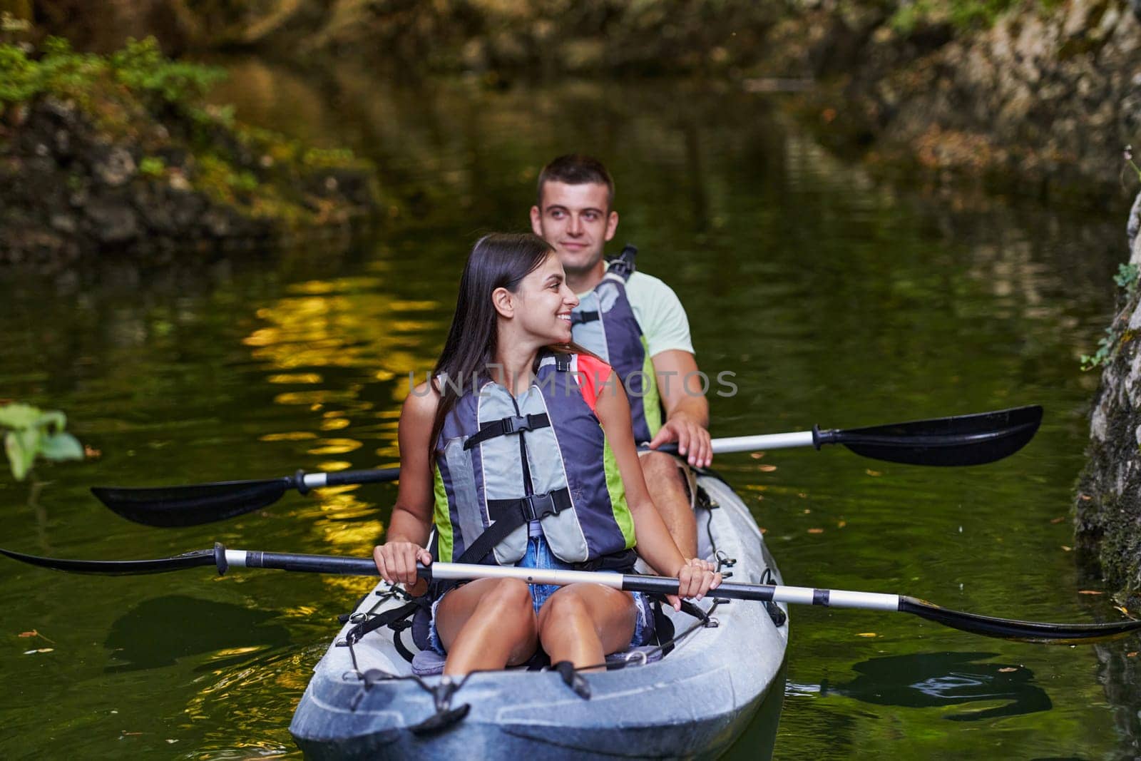 A young couple enjoying an idyllic kayak ride in the middle of a beautiful river surrounded by forest greenery by dotshock