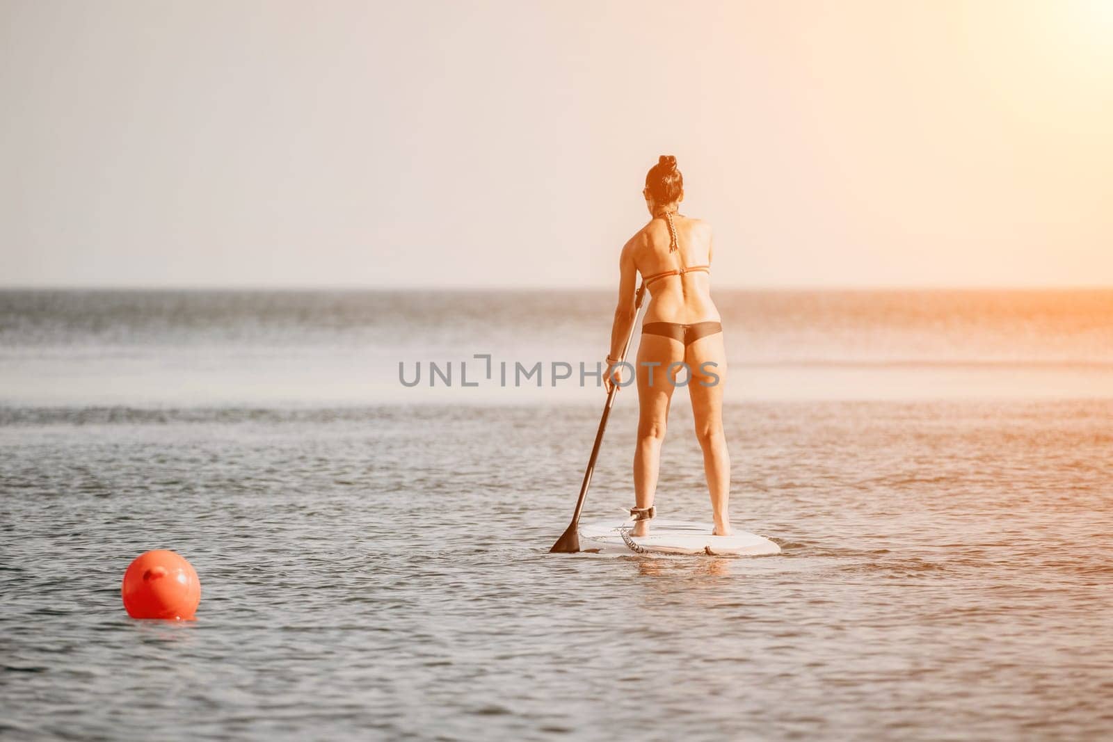 Sea woman sup. Silhouette of happy middle aged tanned woman in rainbow bikini, surfing on SUP board, confident paddling through water surface. Idyllic sunset. Active lifestyle at sea or river. by panophotograph