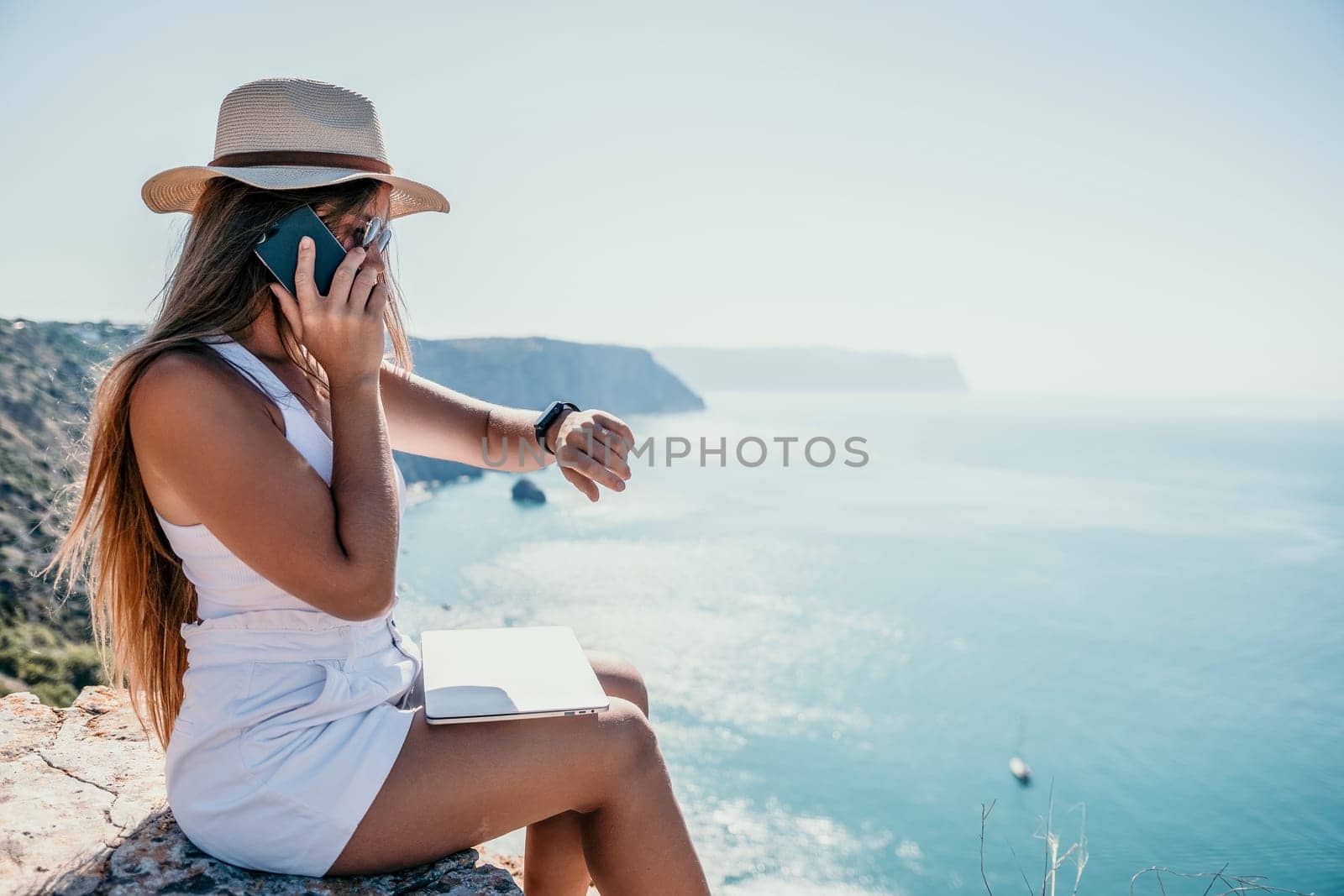 Digital nomad, woman in the hat, a business woman with a laptop sits on the rocks by the sea during sunset, makes a business transaction online from a distance. Freelance, remote work on vacation. by panophotograph