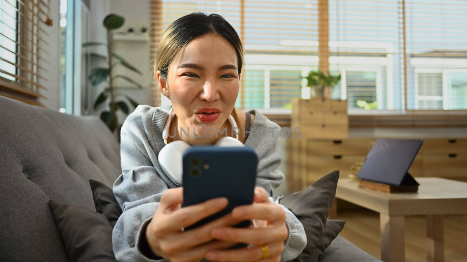 Cheerful young woman lying on sofa in living room using smartphone, texting messaging, communication in social media.