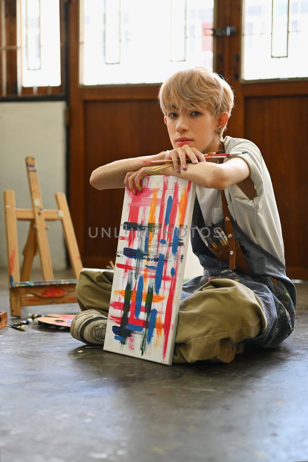 Full length view of young gay male artist sitting on the floor in art studio lit by sunlight.