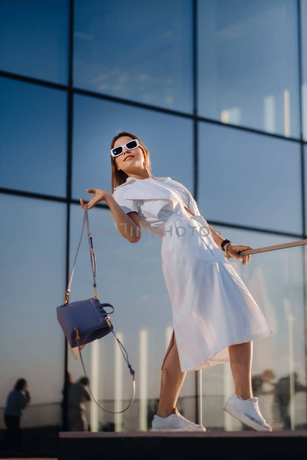 Stylish girl in glasses and a white summer coat in the city with a handbag.