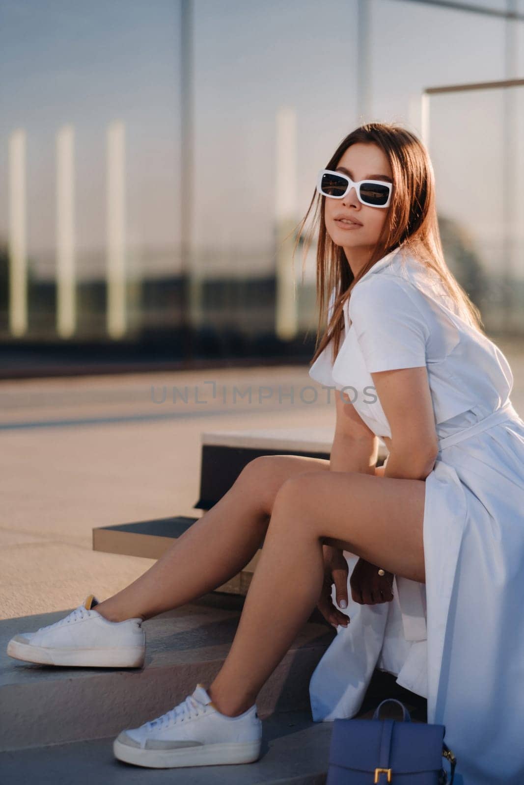 A stylish girl in glasses and a white summer coat is sitting in the city.