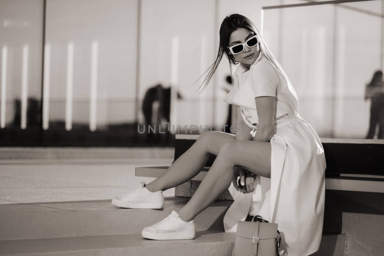 A stylish girl in glasses and a white summer coat is sitting in the city.