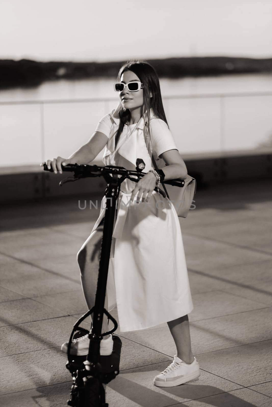 Stylish girl in glasses and a white summer coat in the city on an electric scooter.