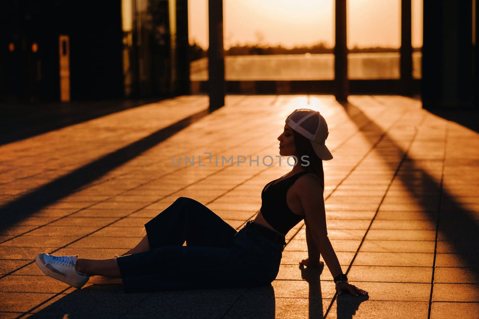 The silhouette of a young teenage girl in jeans and a cap is sitting on the asphalt at sunset.