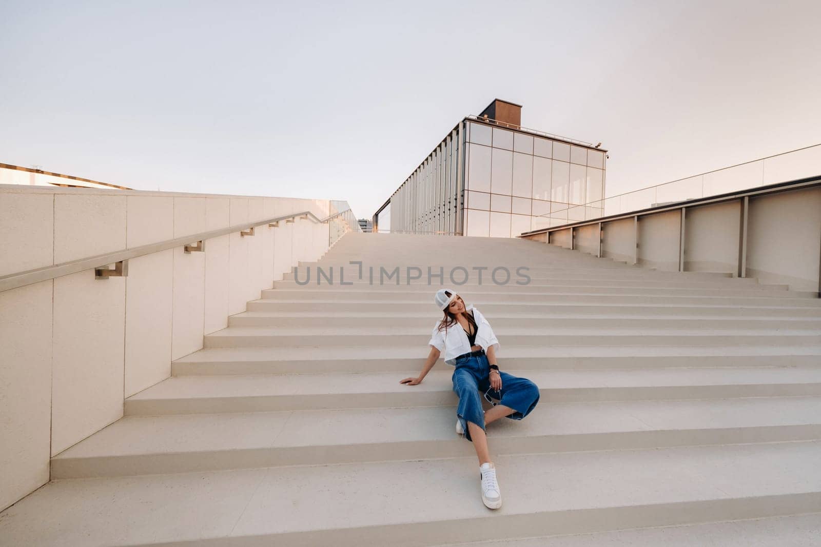 A young teenage girl in jeans and a cap is sitting on the stairs in the city by Lobachad