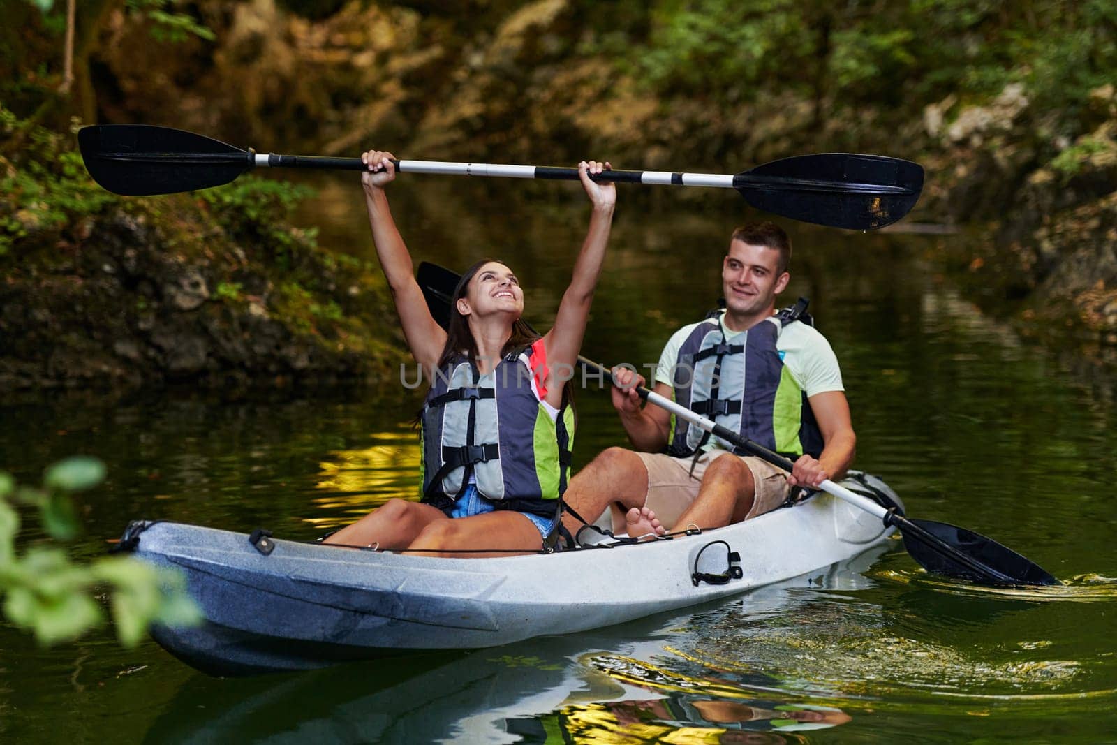 A young couple enjoying an idyllic kayak ride in the middle of a beautiful river surrounded by forest greenery by dotshock