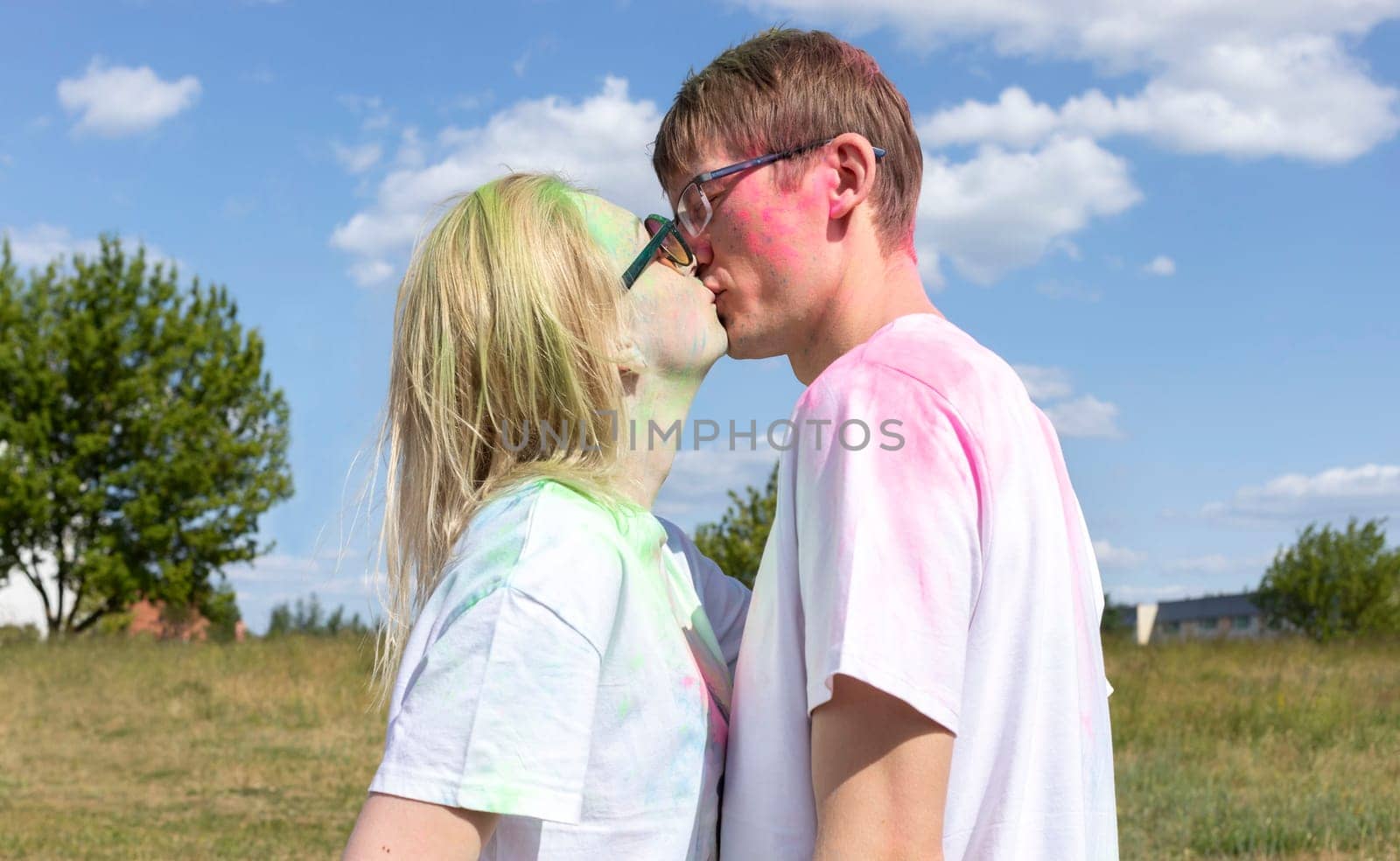 Romantic Caucasian Couple Adults Kiss With Colorful Paint, Powder On Clothes On Holi Color Festival. Emotional Happy Man and Woman. Blue Sky, Green Trees On Background, Sunny Day. Horizontal Plane.