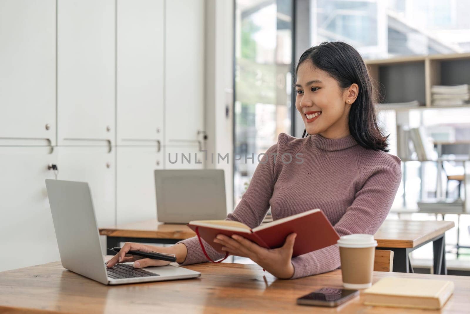 Asian student girl sitting at table looking book while do homework with laptop using internet communicate with friend connection, happy mood smiling broadly in library.