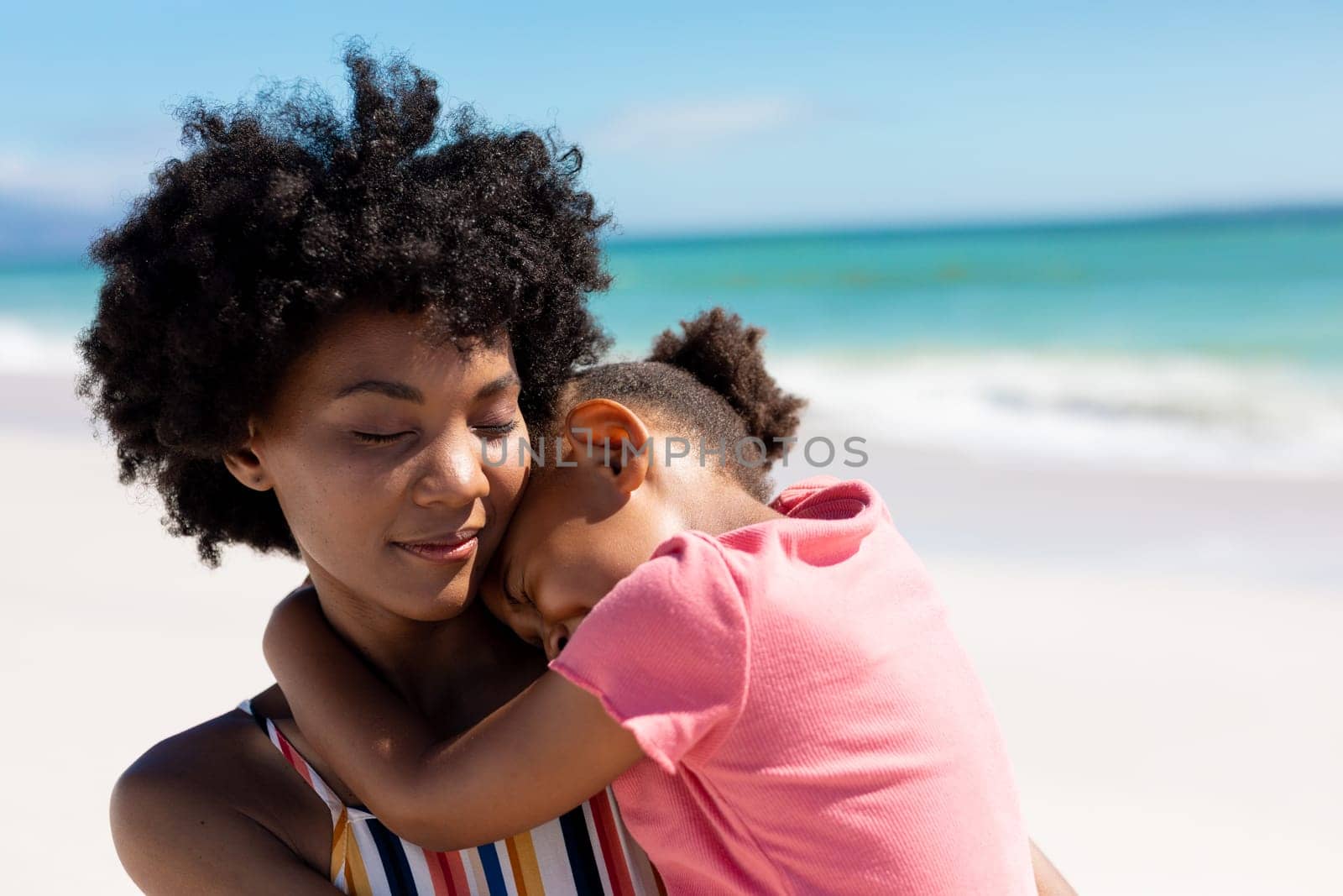 African american woman with eyes closed carrying daughter at beach on sunny day by Wavebreakmedia