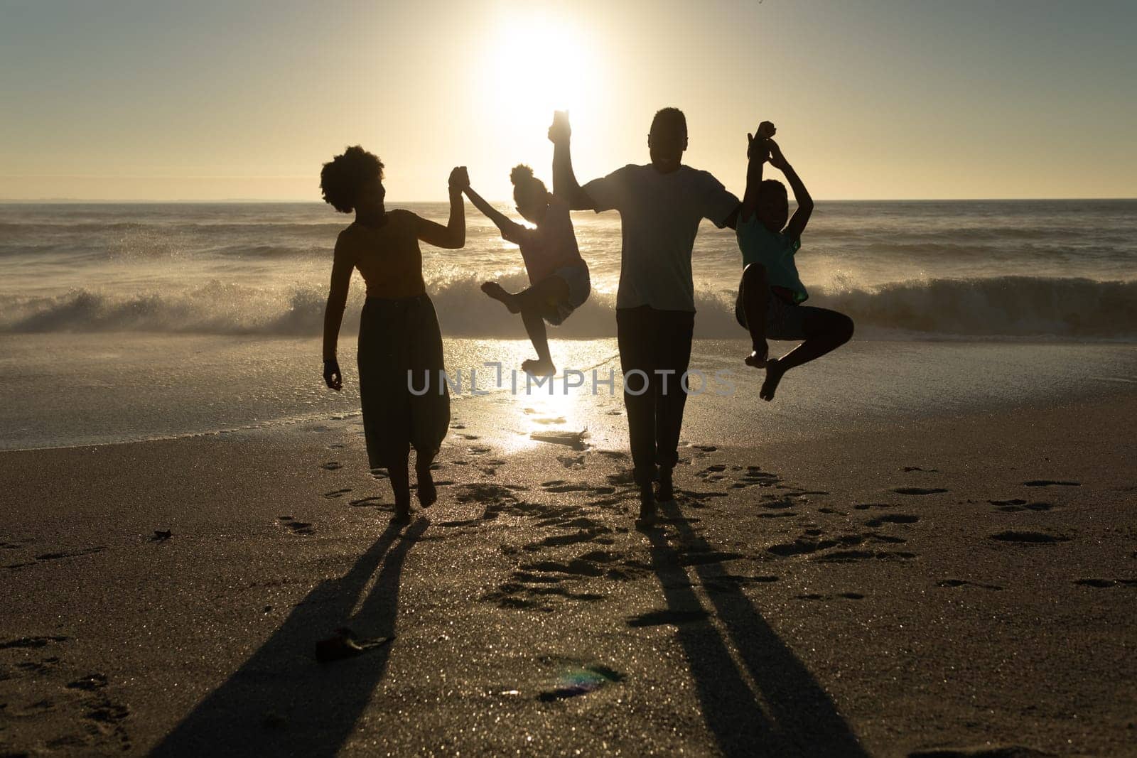 Playful african american parents holding hands of children at beach against sky during sunset. unaltered, family, lifestyle, togetherness, enjoyment and holiday concept.