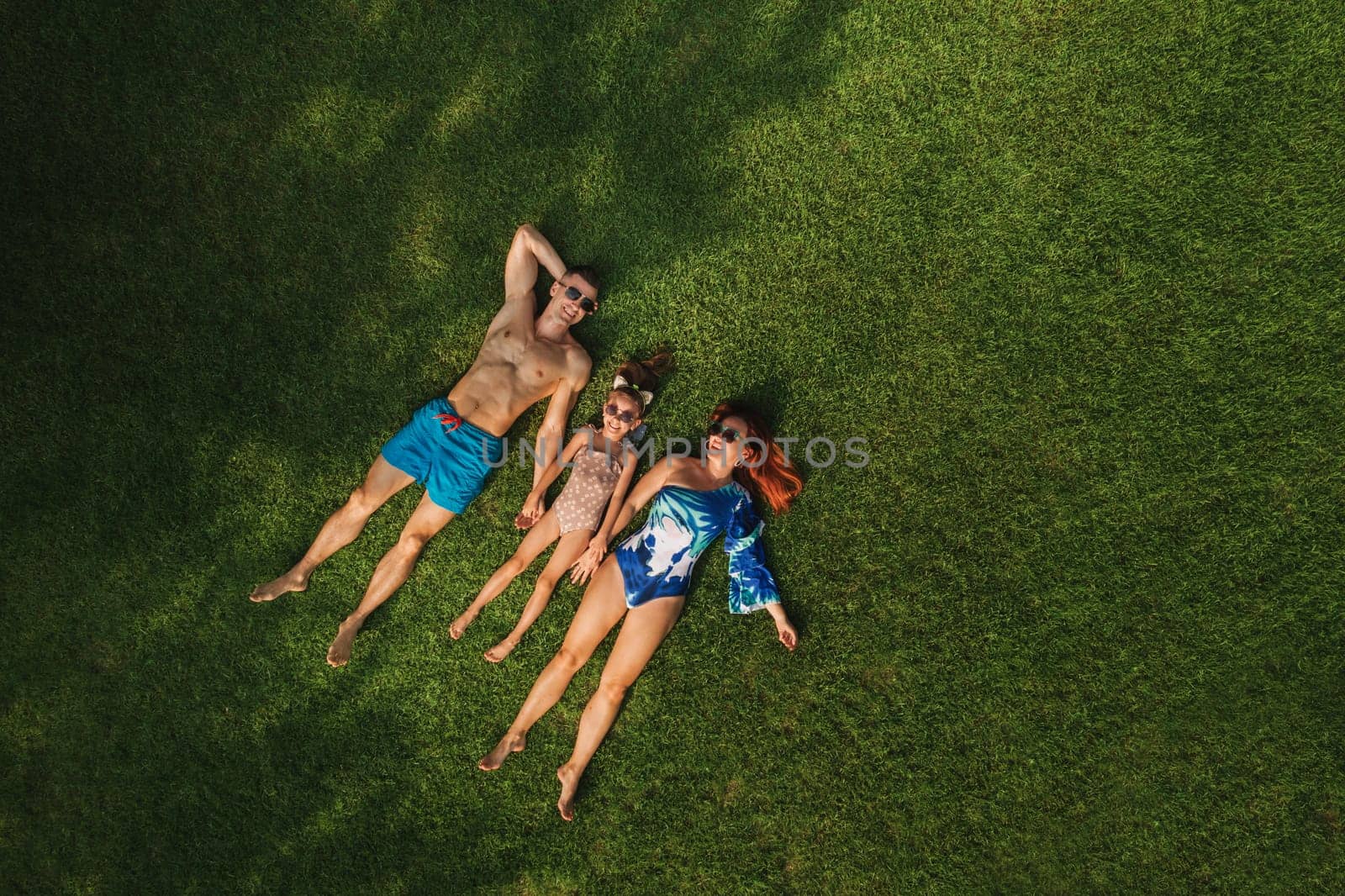Top view of a happy family in swimsuits lying on the green grass by Lobachad
