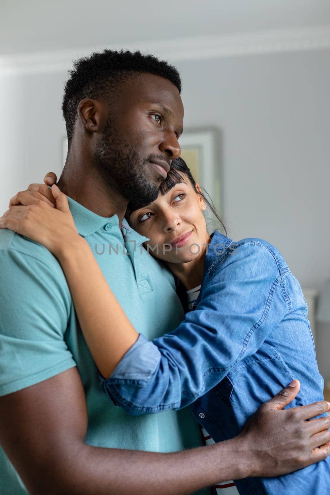 Thoughtful multiracial young couple looking away while embracing in living room at home. unaltered, lifestyle, domestic life, togetherness, love, contemplation.