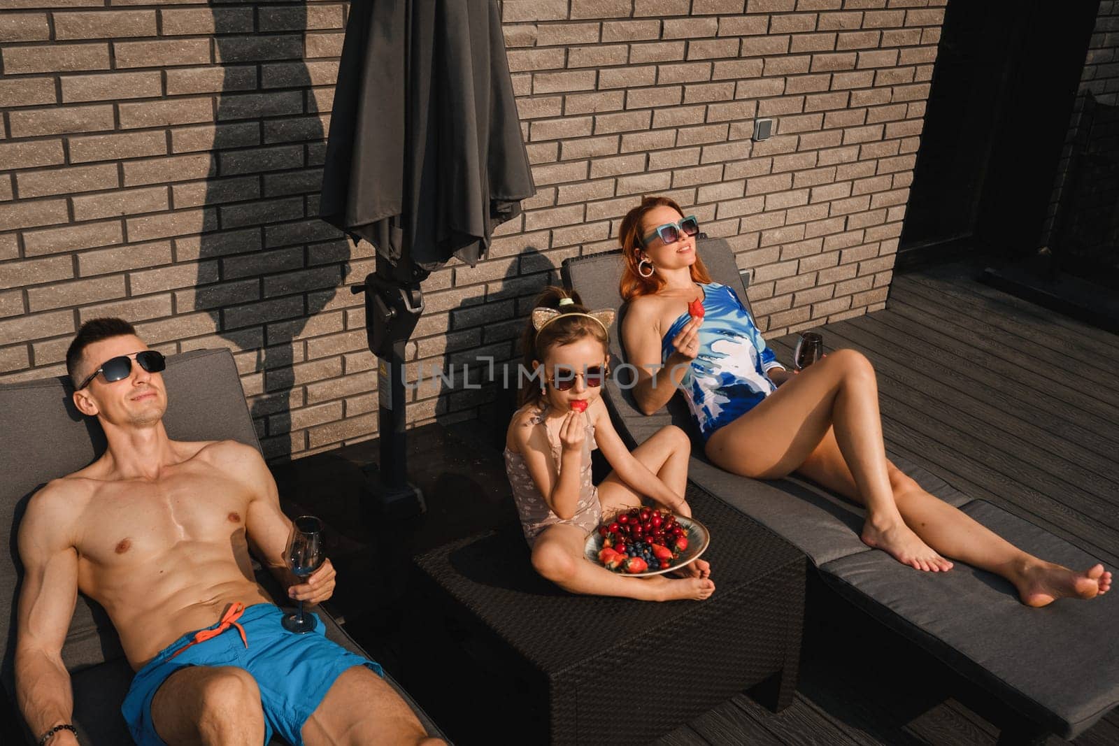 A happy family in swimsuits sunbathe in summer on their terrace on sun beds by Lobachad