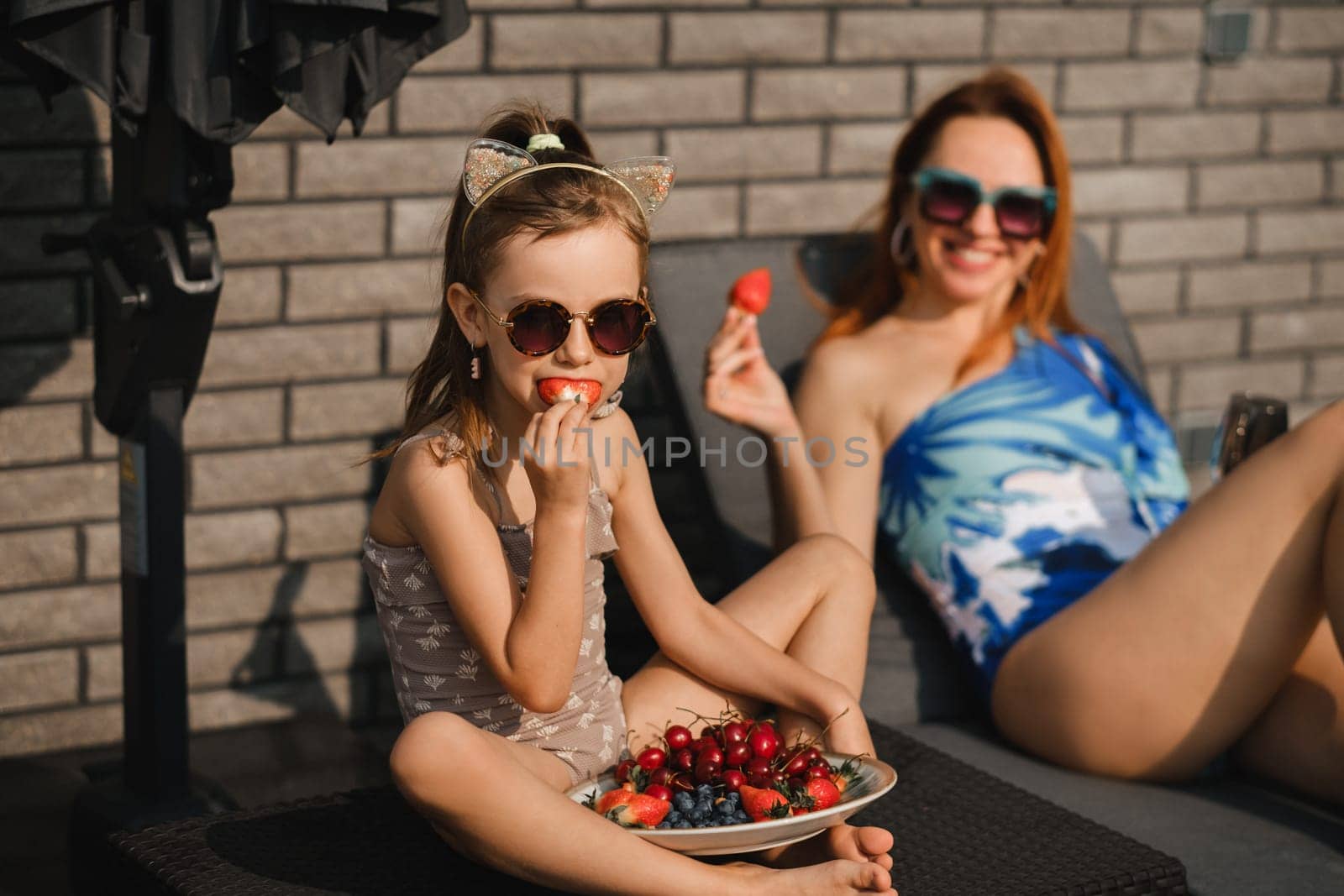 Mom and daughter in swimsuits sunbathe in the summer on their terrace on sun loungers and eats strawberries.