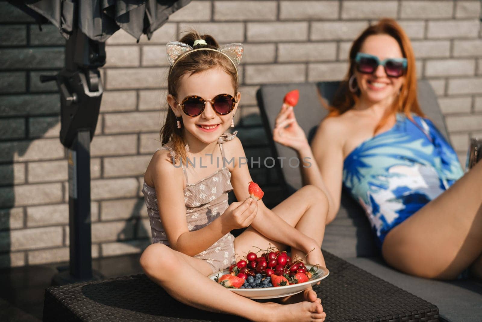 Mom and daughter in swimsuits sunbathe in the summer on their terrace on sun loungers and eats strawberries.