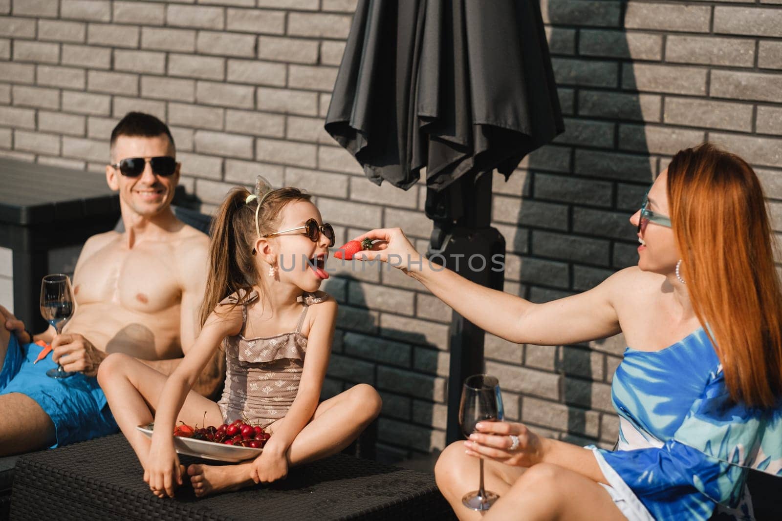 A happy family in swimsuits sunbathes on their terrace in summer. Mom feeds her daughter strawberries.