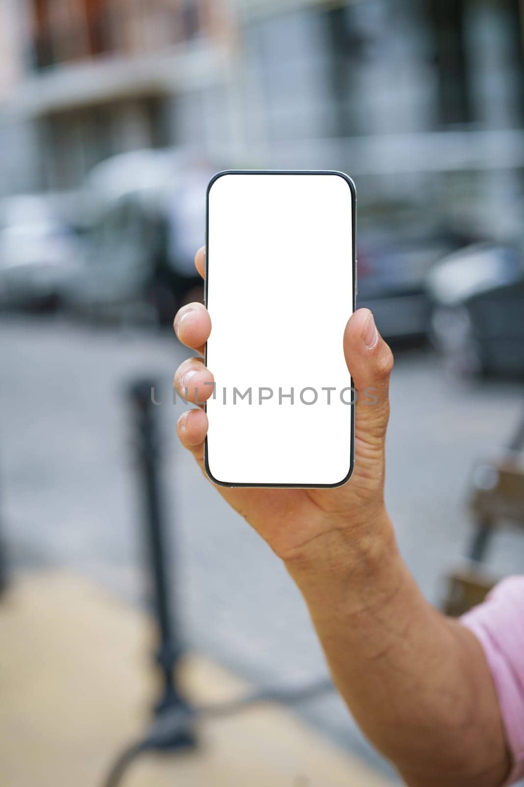 Close-up of man's hand holding mobile phone with white screen display, offering ample copy space. Background features urban cityscape, providing modern and vibrant backdrop. by LipikStockMedia