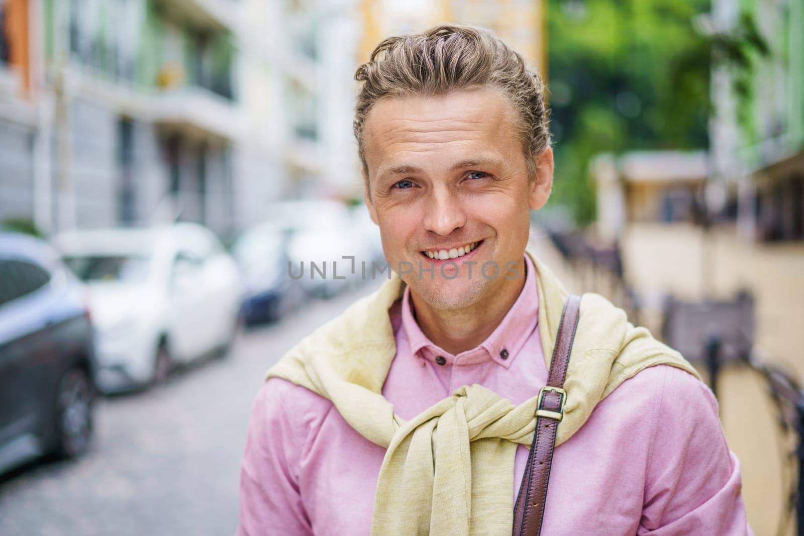 Handsome blond Caucasian man in an old city in Europe. With his charming smile and captivating blue eyes, he gazes directly at the camera, exuding confidence and positivity. by LipikStockMedia