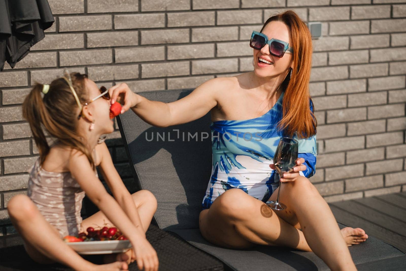A happy family in swimsuits sunbathes on their terrace in summer. Mom feeds her daughter strawberries.