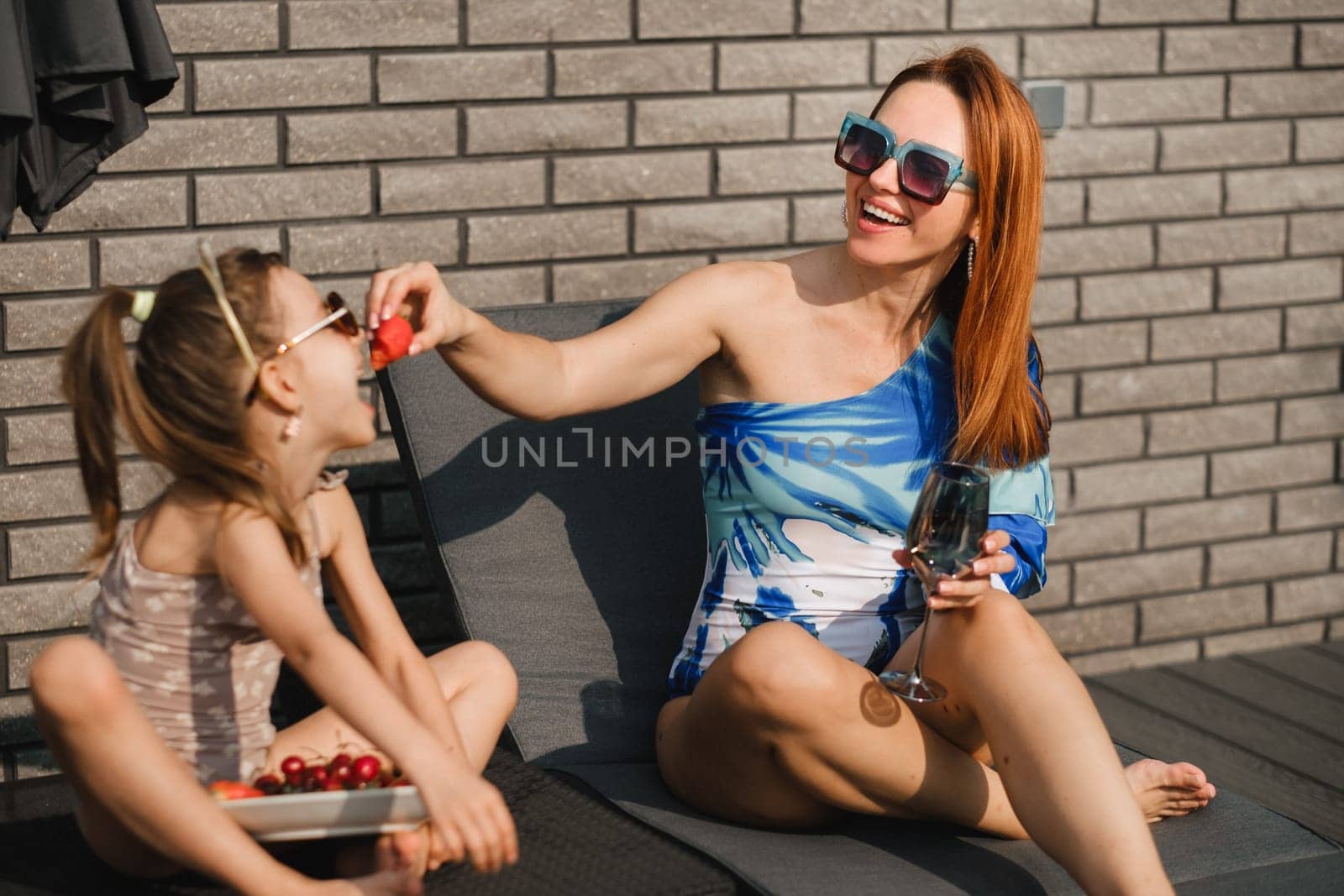 A happy family in swimsuits sunbathes on their terrace in summer. Mom feeds her daughter strawberries.