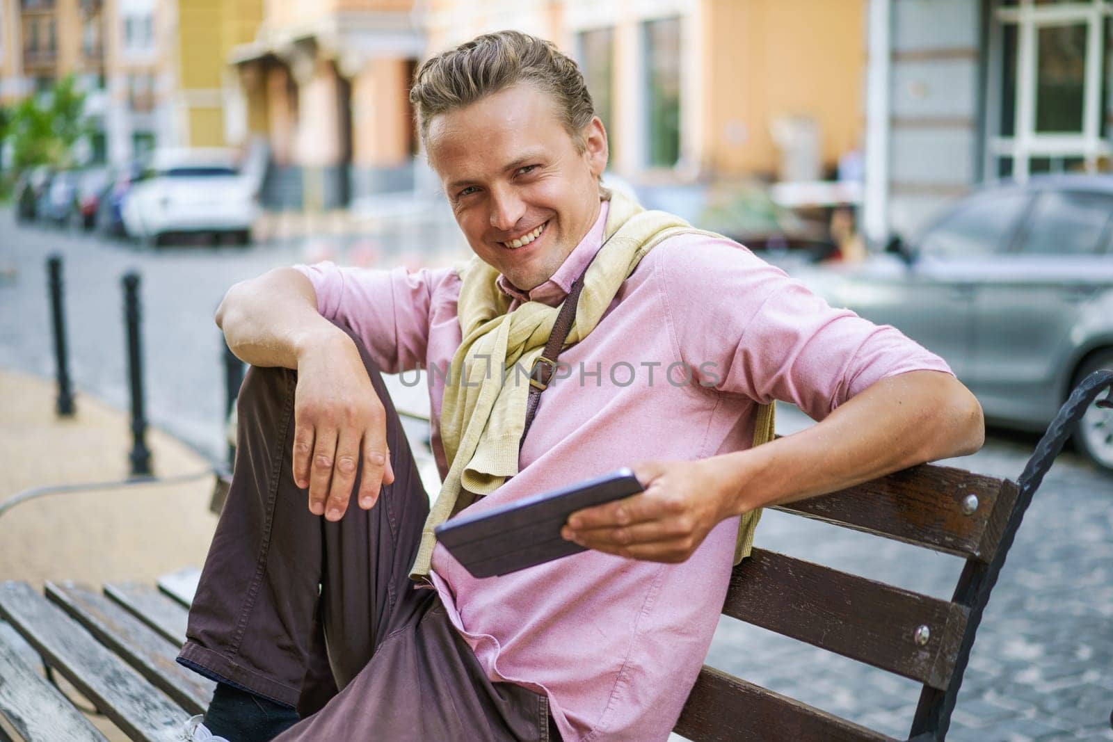 Smiling man sitting on bench in city, and using tablet PC. With gadget in hand, he effortlessly stays connected to digital world, browsing internet, engaging social media, or accessing various digital content. . High quality photo