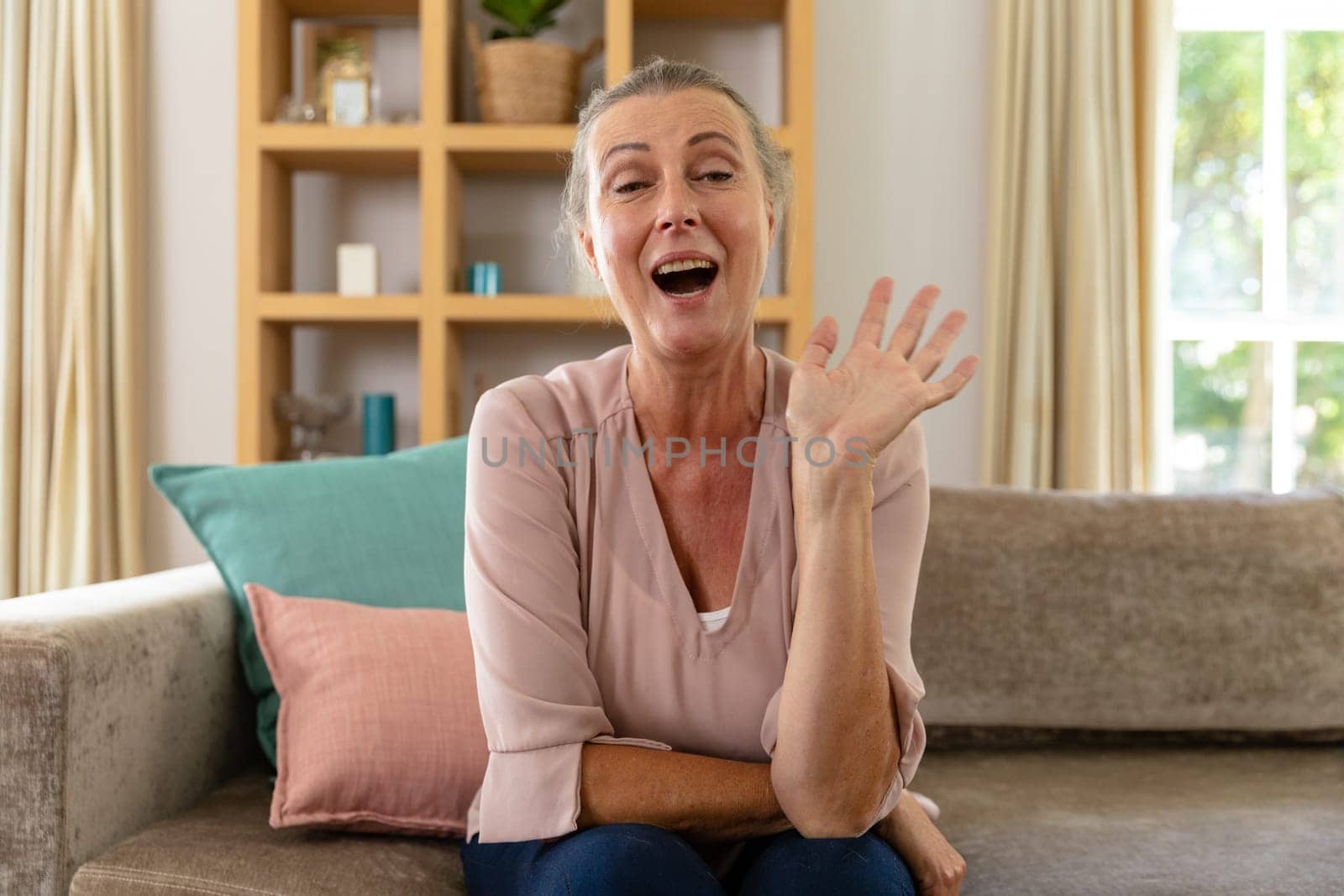 Caucasian senior woman laughing and waving hand while sitting on sofa at home by Wavebreakmedia