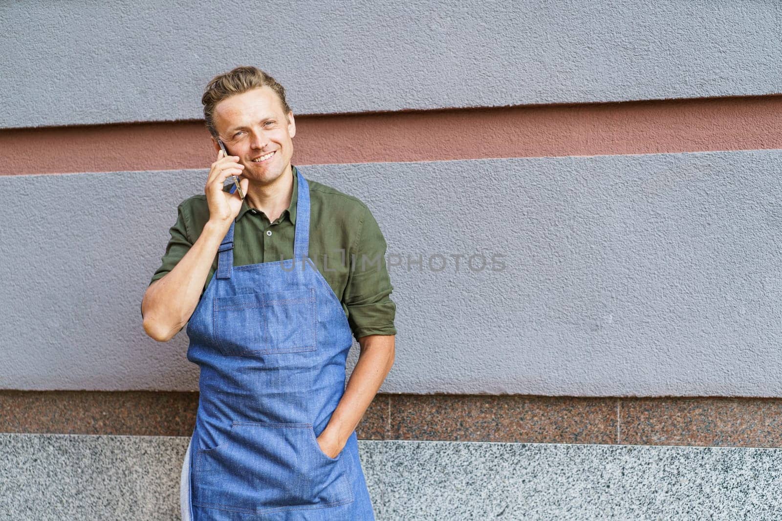 Concept of service readiness and the willingness to assist customers. Worker in blue apron, holding mobile phone, and ready to take order from client against wall background. by LipikStockMedia