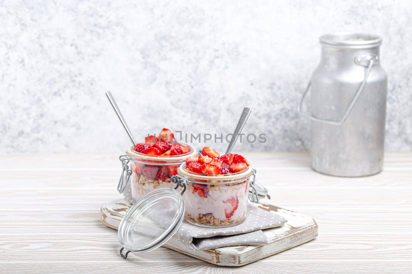 Parfait with Strawberries, Yoghurt and Granola in Transparent Glass Mason Jars on White Rustic Wooden Background from Angle View and Aluminium Milk Can, Healthy Breakfast or Summer Dessert, Copy Space