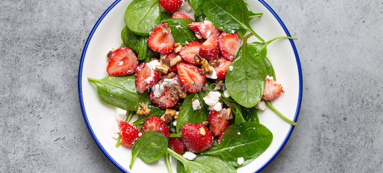 Light Healthy Summer Salad with fresh Strawberries, Spinach, Cream Cheese and Walnuts on White Ceramic Plate, Grey rustic stone Background Top View.