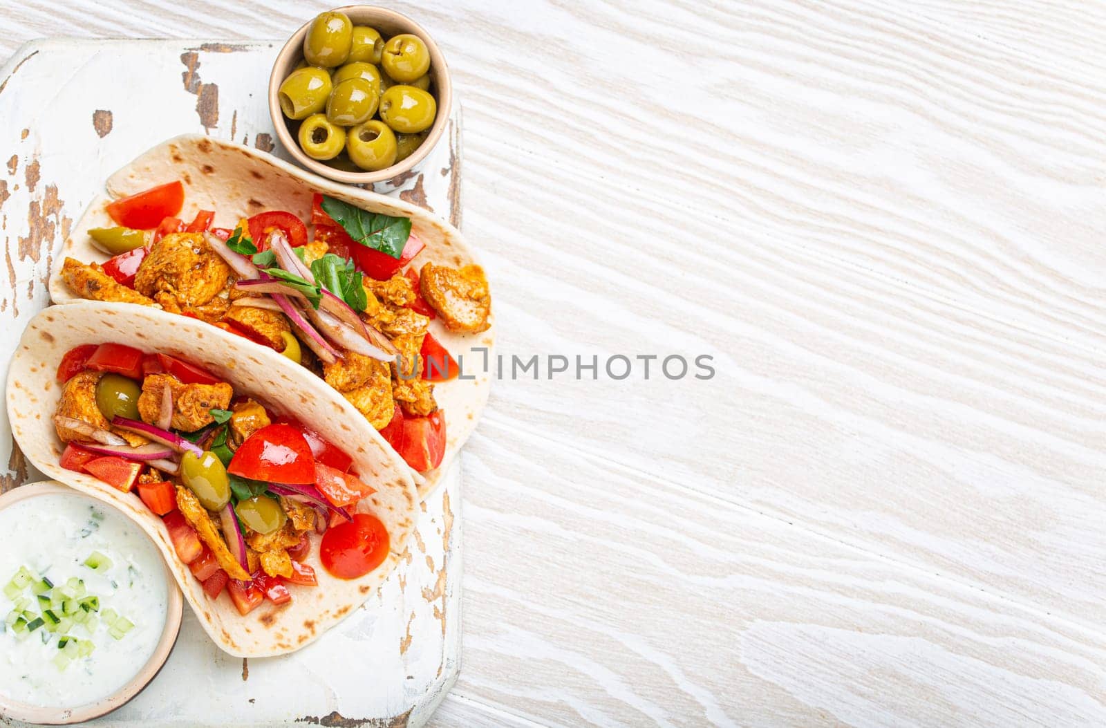 Traditional Greek Dish Gyros: Pita bread Wraps with vegetables, meat, herbs, olives on rustic wooden cutting board with Tzatziki sauce, olive oil top view on white wooden background, space for text.