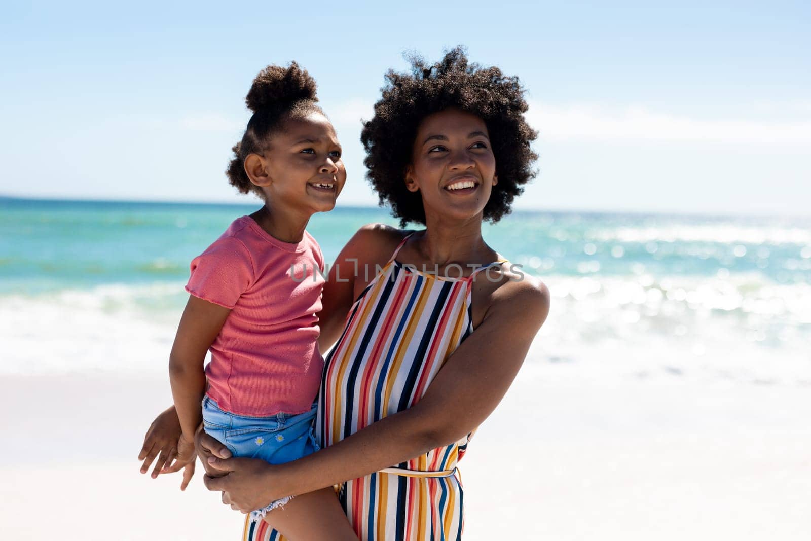 Happy african american mother carrying daughter while enjoying summer weekend at beach against sky. unaltered, family, lifestyle, togetherness, enjoyment and holiday concept.