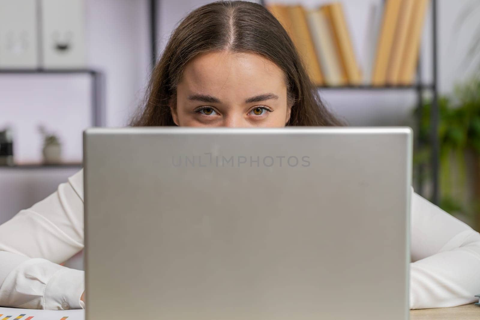 Confident business woman hiding behind laptop computer, looking at camera, spying his colleagues working, peeping. Professional freelancer girl looking from behind computer with cunning eyes glance