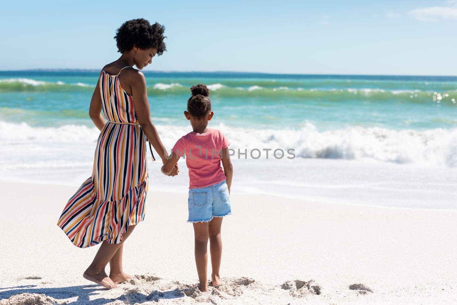 Full length rear view of african american mother and daughter holding hands while standing at beach. unaltered, family, lifestyle, togetherness, enjoyment and holiday concept.