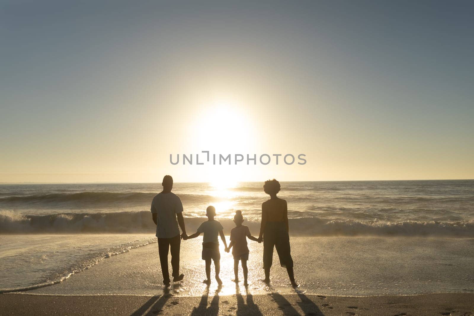 Full length rear view of african american family holding hands while looking at sunset over sea. unaltered, family, lifestyle, togetherness, enjoyment and holiday concept.