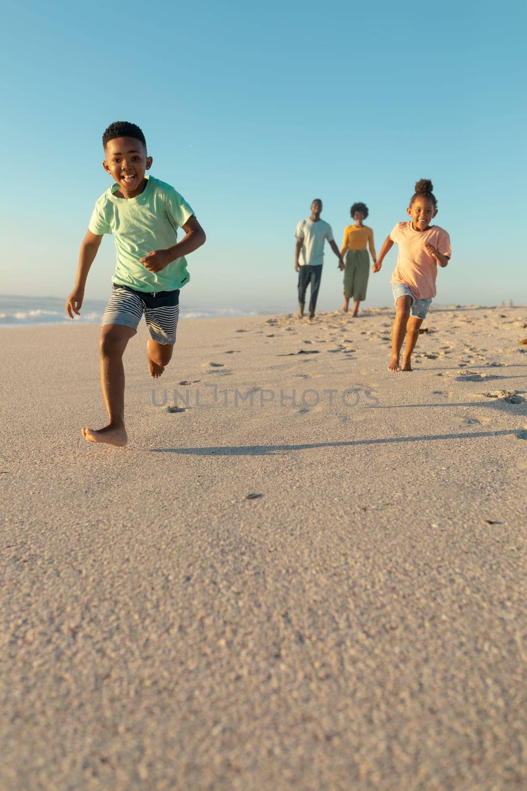 Happy african american siblings running against parents walking at beach on sunny day by Wavebreakmedia