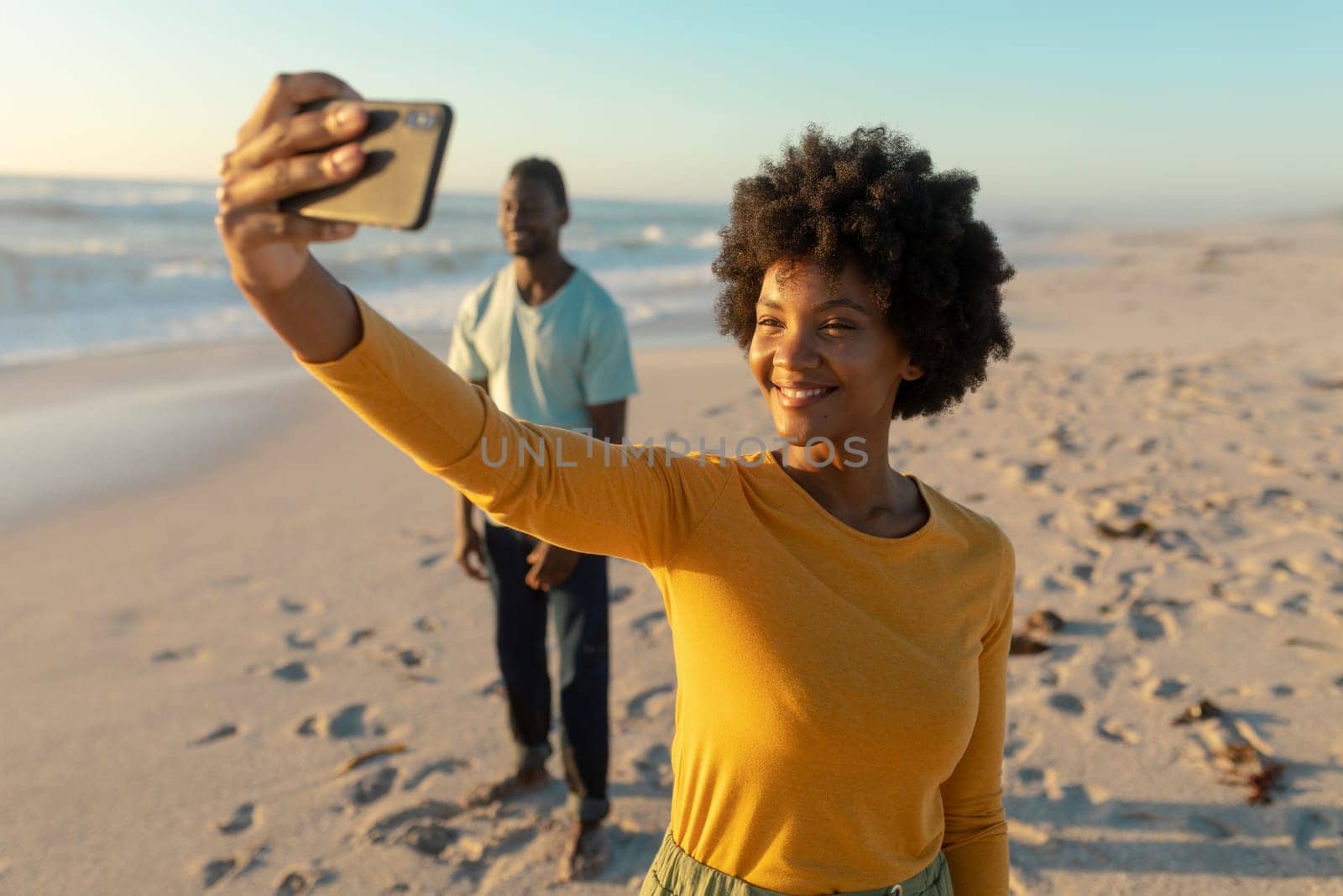 Happy african american woman taking selfie with man standing in background at beach by Wavebreakmedia