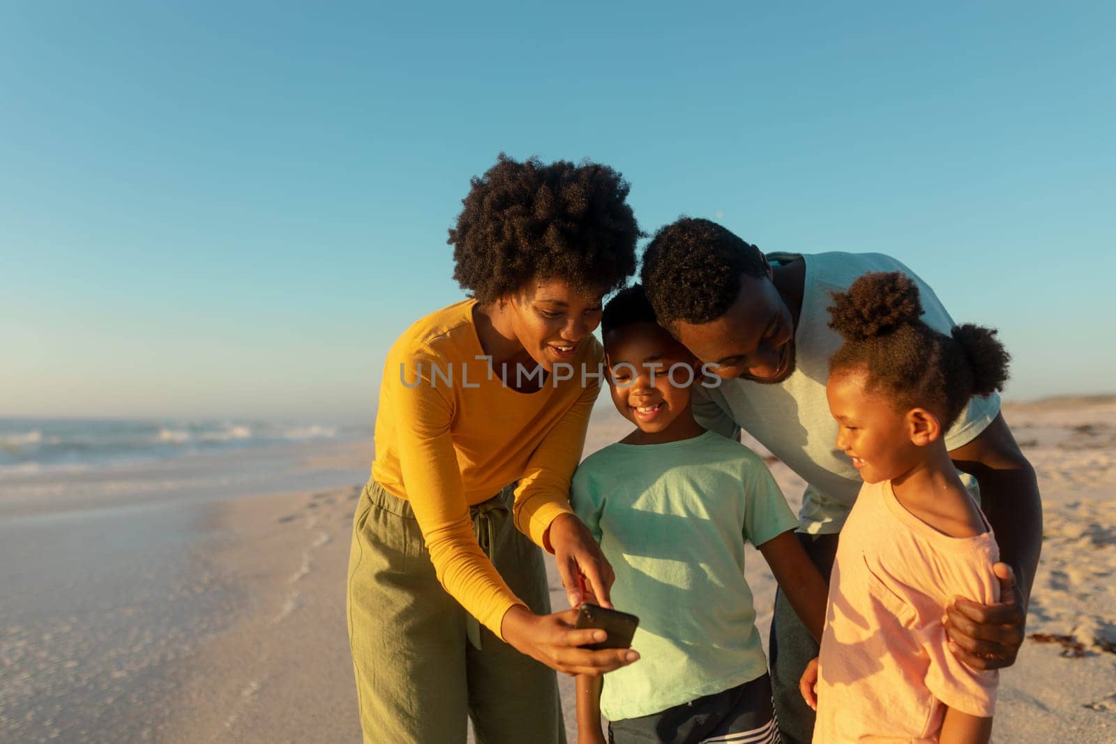 Happy african american mother sharing smartphone with family at beach on sunny day by Wavebreakmedia